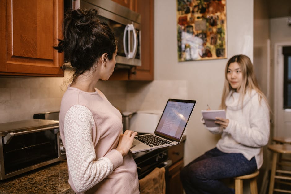 Two women collaborate on laptops and notes in a cozy home kitchen setting